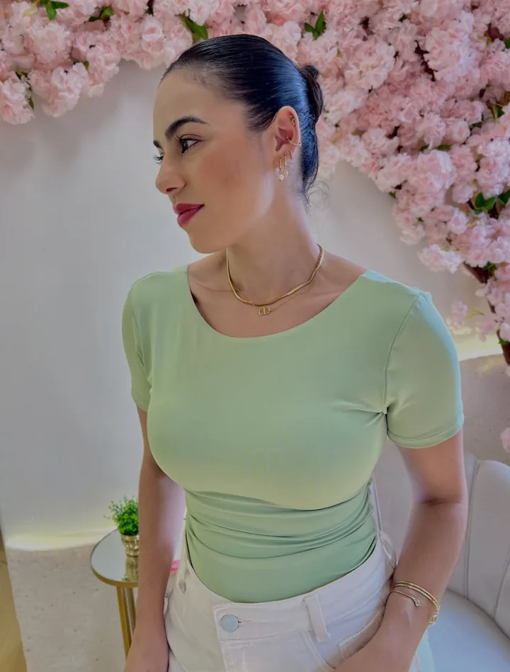 Modelo de perfil com blusa verde de manga curta e calça branca diante de parede de flores