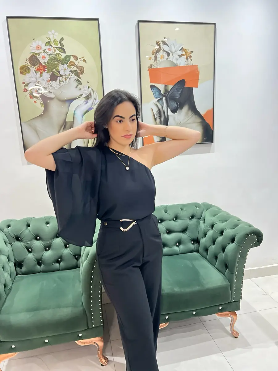 Modelo com look preto de um ombro só, em pé diante de um sofá verde